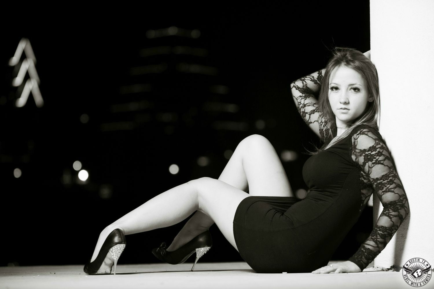 black and white image of a sandy haired fashion model in a tight short black dress with black heels with bling and lots of leg showing at the long center with the austin skyline at night in the background